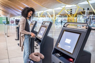 happy black woman using the check-in machine at the airport getting the boarding pass. clipart