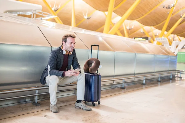 젊은 남자 대기 듣는 음악 및 가방을 공항에서 휴대 전화를 사용 하 여 — 스톡 사진