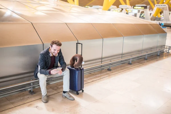Jovem esperando ouvir música e usar o telefone celular no aeroporto com uma mala — Fotografia de Stock