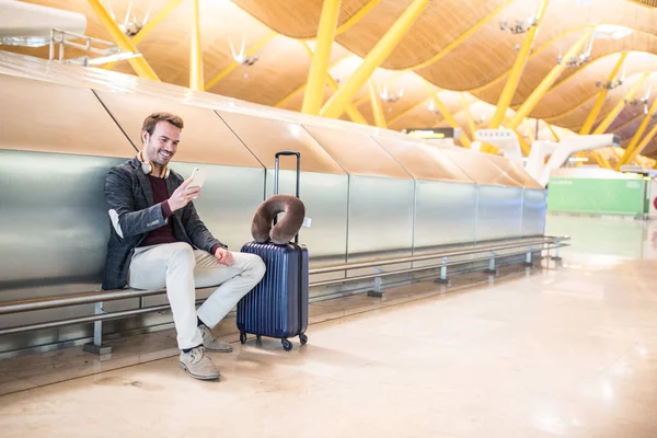 Joven esperando escuchando música y usando un teléfono móvil en el aeropuerto con una maleta — Foto de Stock