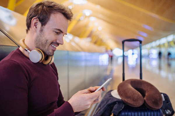 Young man waiting listening music and using mobile phone at the airport with a suitcase