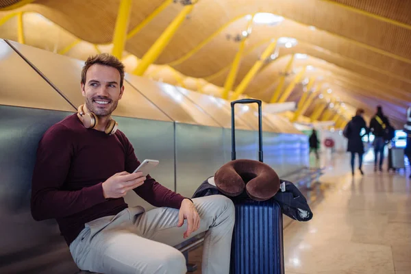 Young man waiting listening music and using mobile phone at the airport with a suitcase — Stock Photo, Image