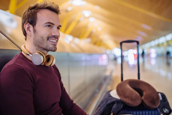 Młody człowiek czeka słuchania muzyki i przy użyciu telefonu komórkowego na lotnisku z walizką — Zdjęcie stockowe