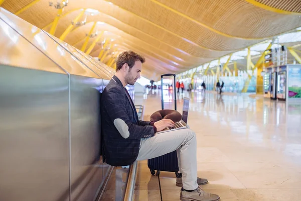 Jovem homem atraente sentado no aeroporto trabalhando em seu laptop esperando seu voo com uma mala . — Fotografia de Stock