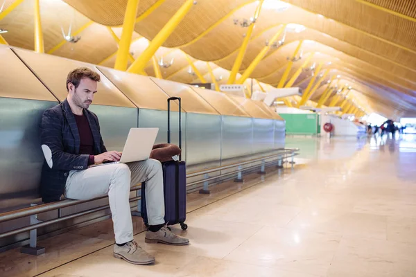Young attractive man sitting at the airport working in his laptop waiting his flight with a suitcase. — Stock Photo, Image