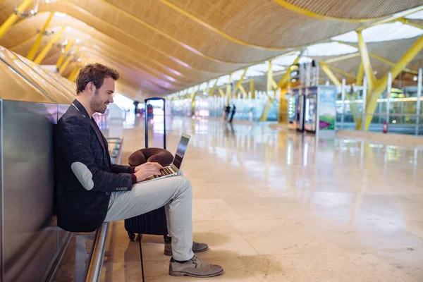 Young attractive man sitting at the airport working in his laptop waiting his flight with a suitcase. — Stock Photo, Image