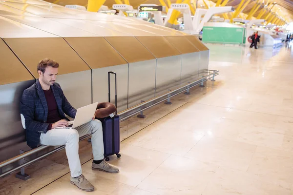 Jeune homme séduisant assis à l'aéroport travaillant dans son ordinateur portable attendant son vol avec une valise . — Photo