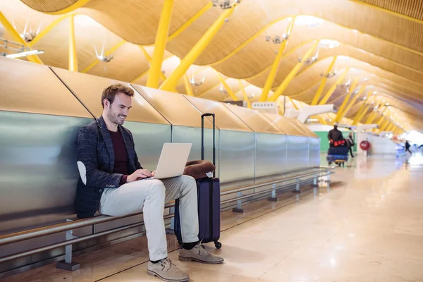 Young attractive man sitting at the airport working in his laptop waiting his flight with a suitcase. — Stock Photo, Image