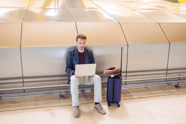 Joven hombre atractivo sentado en el aeropuerto trabajando en su portátil esperando su vuelo con una maleta . — Foto de Stock