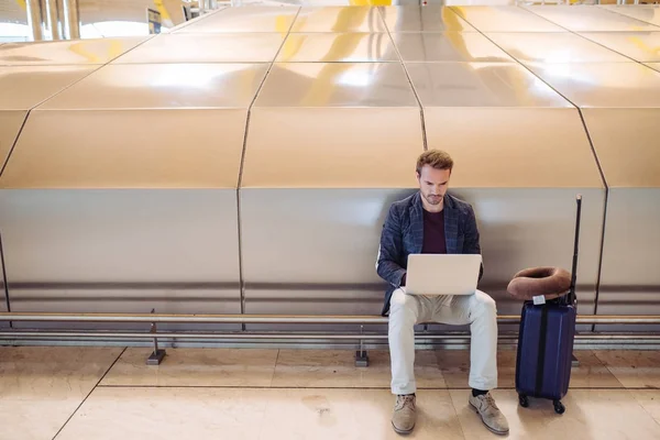 Młody atrakcyjny człowiek siedzi na lotnisku działa w swoim laptopie czeka jego lotu z walizką. — Zdjęcie stockowe