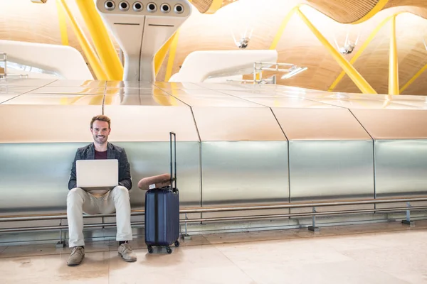 Jeune homme séduisant assis à l'aéroport travaillant dans son ordinateur portable attendant son vol avec une valise . — Photo