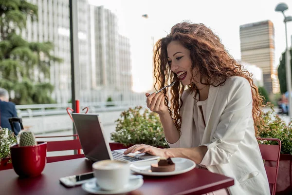 Молодая деловая рыжая женщина за компьютером и ест торт — стоковое фото