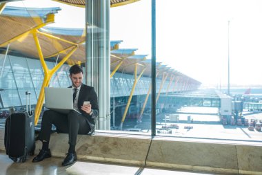 genç yakışıklı işadamı onun dizüstü bilgisayar ve cep telefonu havaalanında uçağı için bekleyen ve gülümseyen kullanarak