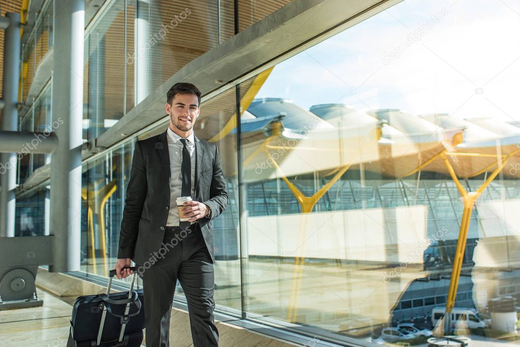 young businessman walking at the airport terminal with luggage smiling with a coffee 