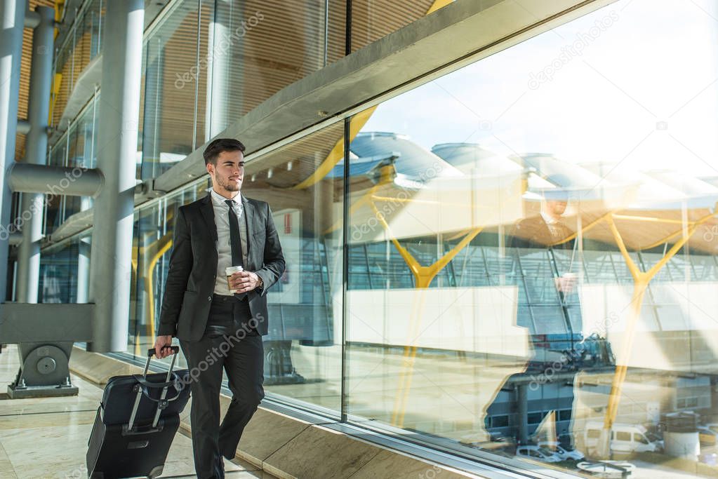 young businessman walking at the airport terminal with luggage smiling with a coffee 