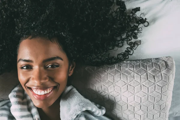Черная женщина с кудрявыми волосами улыбается и лежит на кровати, глядя в камеру — стоковое фото