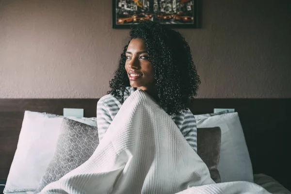 Όμορφη μαύρη γυναίκα που καλύπτεται με φύλλο και κάθονται χαμογελαστή στο κρεβάτι που βλέπει μακριά — Φωτογραφία Αρχείου