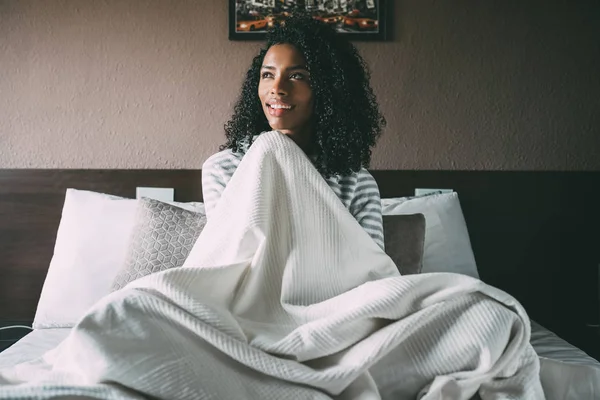 Гарненька чорна жінка вкрита простирадлом і сидить на ліжку, дивлячись вниз — стокове фото