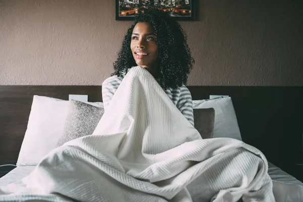 Bonita mujer negra cubierta con sábanas y sentada en la cama mirando hacia otro lado — Foto de Stock