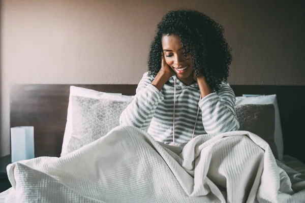 Bonita mujer negra cubierta con sábanas y sentada en la cama mirando hacia otro lado — Foto de Stock