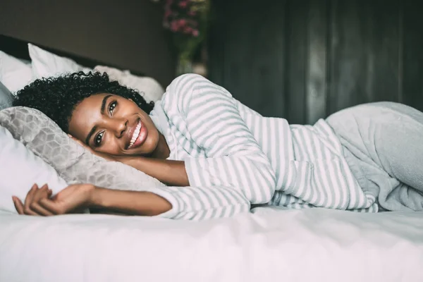 Close up de uma mulher negra bonita com cabelo encaracolado sorrindo e deitado na cama olhando para longe — Fotografia de Stock