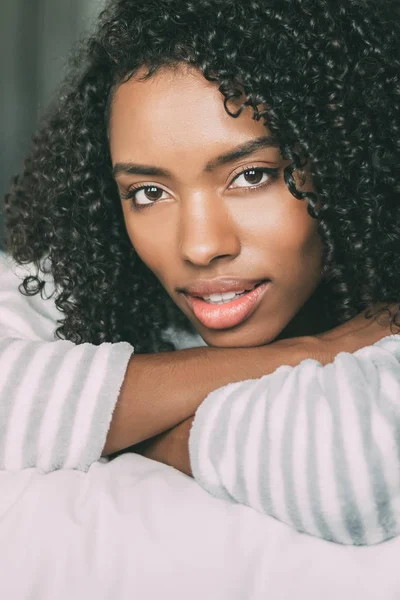 Primer plano de una bonita mujer negra con el pelo rizado sonriendo y acostada en la cama mirando a la cámara —  Fotos de Stock