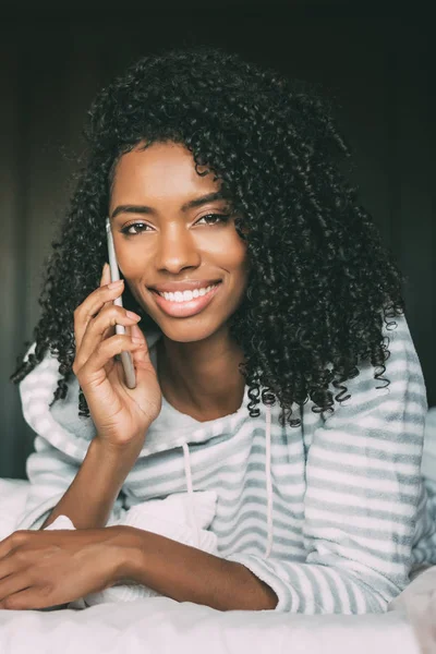 Close up of a pretty black woman with curly hair with smartphone on bed looking at the camera — Stock Photo, Image
