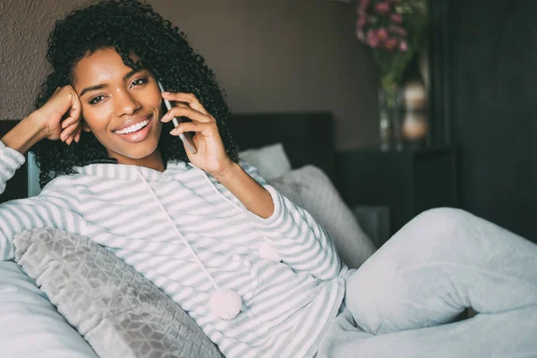 Κοντινό πλάνο του μια όμορφη μαύρη γυναίκα με σγουρά μαλλιά χαμογελώντας και χρήση του τηλεφώνου σε κρεβάτι αναζητούν μακριά — Φωτογραφία Αρχείου