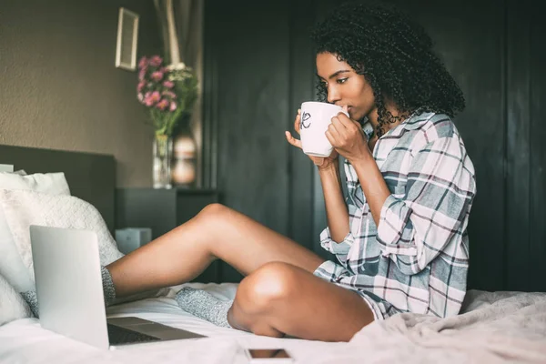 Beautiful black woman on bed with laptop and cup of coffee — Stock Photo, Image