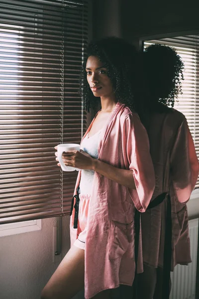 Όμορφη μαύρη γυναίκα που στέκεται στο παράθυρο με φλιτζάνι καφέ — Φωτογραφία Αρχείου