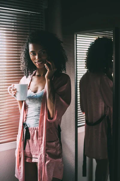 Όμορφη μαύρη γυναίκα που στέκεται στο παράθυρο με το Κύπελλο του καφέ και του smartphone — Φωτογραφία Αρχείου