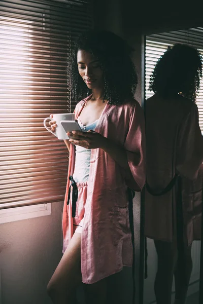 Pretty black woman standing at the window with cup of coffee and smartphone — Stock Photo, Image