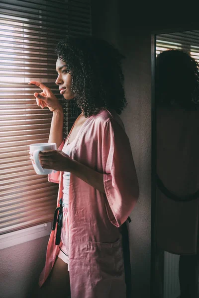 Όμορφη μαύρη γυναίκα που στέκεται στο παράθυρο με φλιτζάνι καφέ — Φωτογραφία Αρχείου