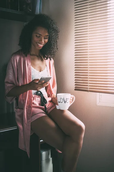 Kaunis musta nainen istuu ikkunassa kupin kahvia ja älypuhelin — kuvapankkivalokuva