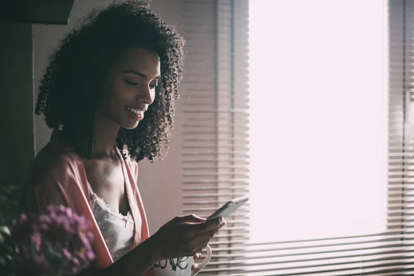 Όμορφη μαύρη γυναίκα κάθεται στο παράθυρο με το Κύπελλο του καφέ και του smartphone — Φωτογραφία Αρχείου