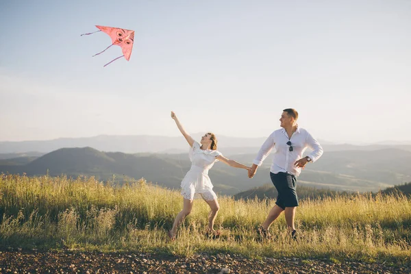 Běhání s létající drak bavte se na venkově. Chlapec a dívka běží na kopci s létajícím drakem proti modré obloze a slunečním paprskům. — Stock fotografie