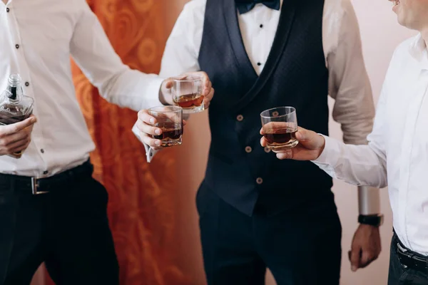 Ženich a jeho kamarádi ve stylových oblecích popíjejí whisky v hotelovém pokoji. Ráno před přípravou svatby. Skupina přátel slaví. Royalty Free Stock Fotografie