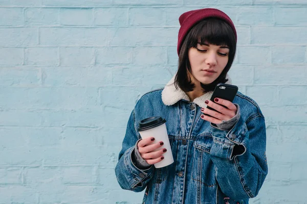 Νεαρό κορίτσι hipster χρησιμοποιεί το τηλέφωνο και κρατάει ένας καφές — Φωτογραφία Αρχείου