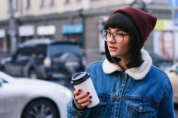 Stylish young hipster girl walks in the city and drinking coffee on the background of cars — Stock Photo, Image