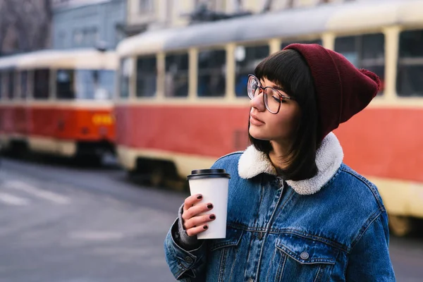 Κομψό νεαρό hipster κορίτσι βόλτες στην πόλη και πίνει καφέ στο φόντο του τραμ — Φωτογραφία Αρχείου