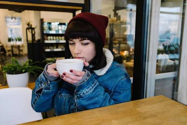 Elegante jovem hipster menina sentado em um café bebendo café e olhando na janela — Fotografia de Stock