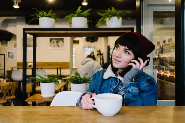 Joven chica hipster feliz hablando por teléfono y bebiendo una bebida caliente en la cafetería — Foto de Stock