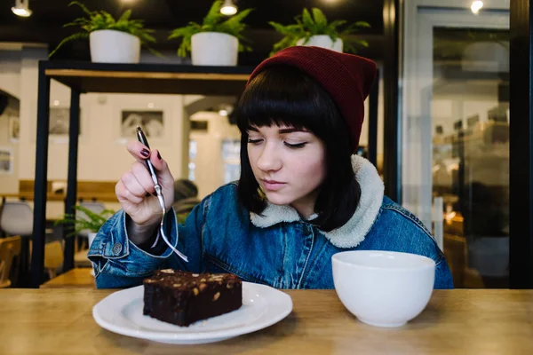 Joven chica hipster come sabroso pastel de chocolate y beber café caliente en un agradable café — Foto de Stock
