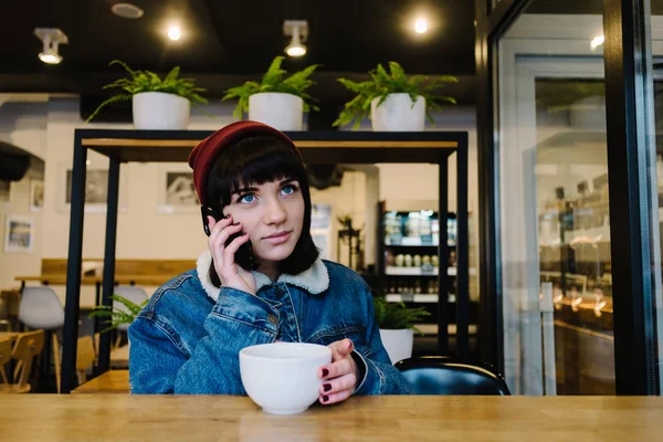 Молодая счастливая хипстерша разговаривает по телефону и пьет чай в красивом кафе — стоковое фото