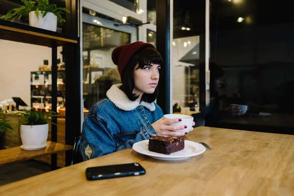 Joven chica hipster come sabroso pastel de chocolate y beber té caliente en un hermoso café — Foto de Stock