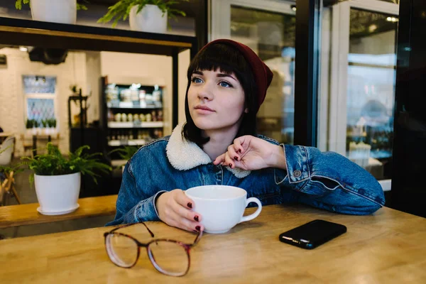 Elegante jovem hipster menina sentado em um café bebendo café e olhando na janela — Fotografia de Stock