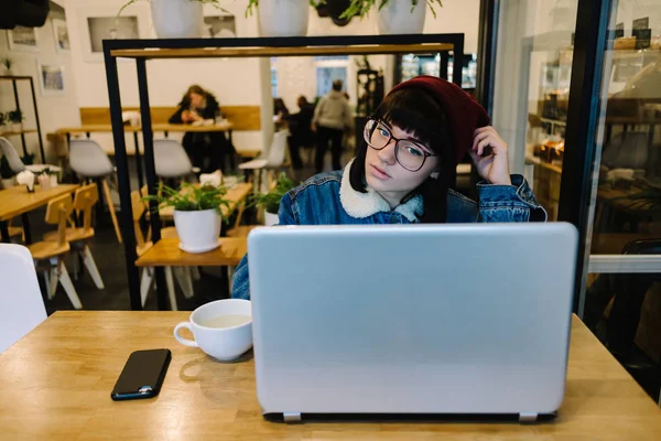 Fiatal lány csípő néz érdeklődéssel a laptop, és kávét iszik egy szép kávézó — Stock Fotó