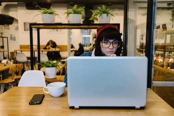 Joven chica hipster mira con interés en su ordenador portátil y beber caliente d bebida en un agradable café —  Fotos de Stock