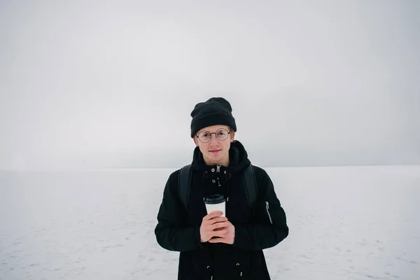 Sonriente joven hipster tipo vestido con gafas de sol negras y el invierno en el lago cubierto de nieve y una taza de café en la mano — Foto de Stock