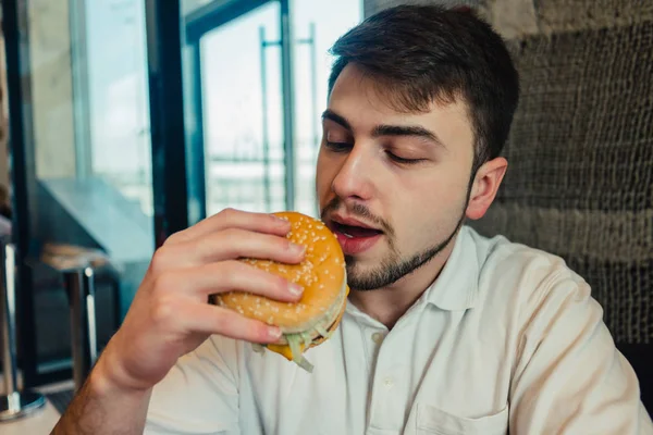 Ein junger Mann sitzt in einem Restaurant und isst leckere Burger — Stockfoto